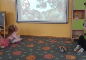 Dzieci z grupy fioletowej siedzą w półkolu na dywanie w sowy i oglądają filmik na temat dymów i ich źródeł na tablicy multimedialnej